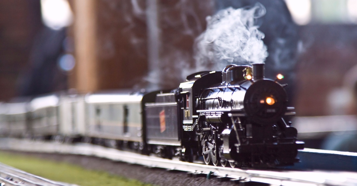 A closeup of a small model steam train pulling multiple cars behind it. A small trail of steam is coming from the lead car.